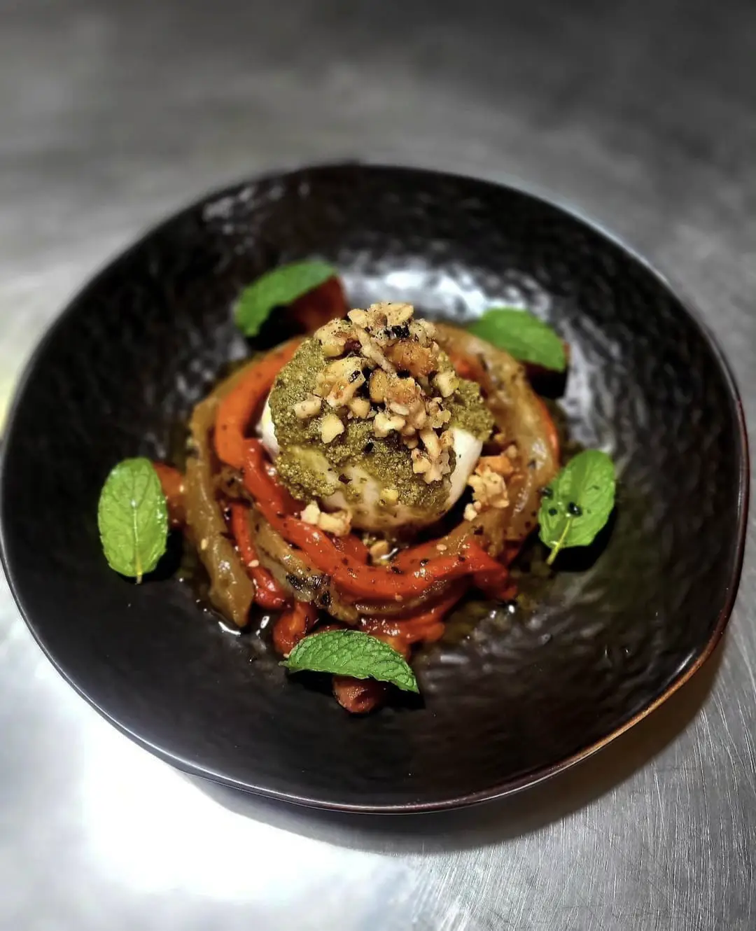 Burrata acompañada de pimientos y berenjena escalivados (asados previamente) a la brasa. Tomate cherry especiado y asado, pesto, frutos secos garrapiñados, menta.